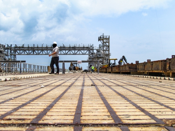 Large-format photograph: Pad Deck, Launch Complex 39-B, showing Apollo Era Crawlerway Grid Panels east of the Flame Trench in the area just south of the Catacombs Roof, where the Panels are divided into two runs, one each for both of the Crawler Trackways. In the distance, ongoing work can be seen, proceeding with the removal of the Grid Panels and refurbishment of the Cutout in the Catacombs Roof where they were removed from. Many of the already-removed Panels can be seen stacked on cribbing in the right side of this image. Photo credit: Withheld by request.